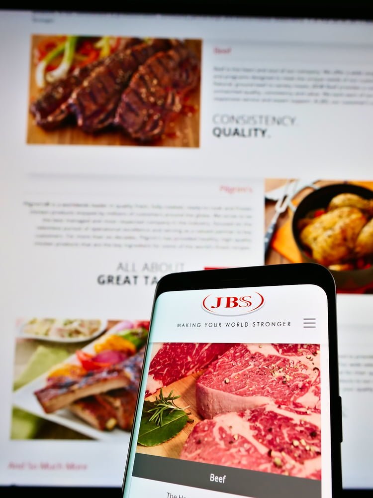 Mobilní telefon s webovými stránkami a logem brazilské společnosti JBS S.A. na pozadí.