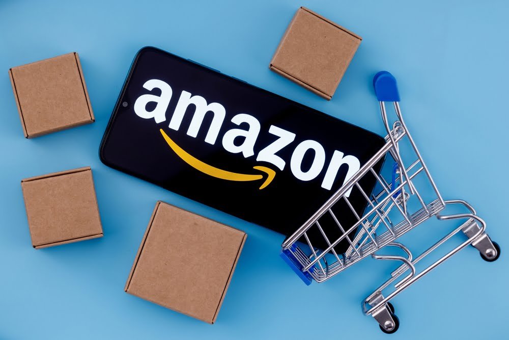 Telefon zobrazující logo Amazonu, papírové boxy a nákupní vozík. Světlé modré pozadí. 