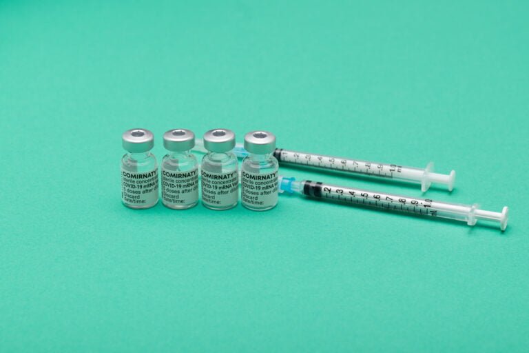 Čtyři ampule vakcíny od Pfizeru proti COVID-19, s jehlami a stříkačkami na zeleném pozadí.