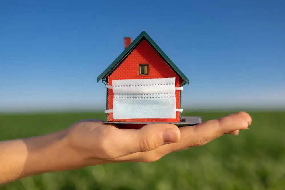 Ruka držící malý model domu s rouškou, v pozadí je zelená louka a modré nebe. Koncept investování do nemovitostí v době pandemie