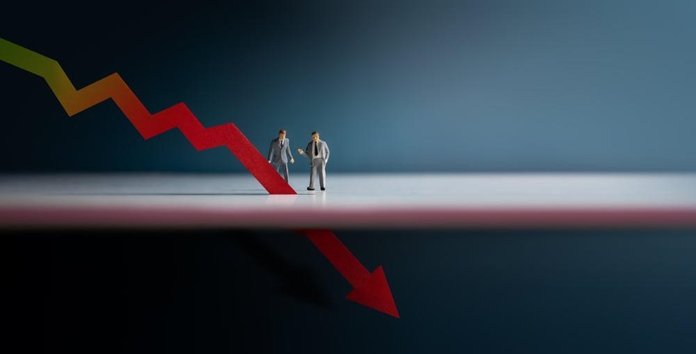 Dvě miniaturní postavičky podnikatele, který se dívají na červenenou šipku klesající směrem dolů. Koncept co je recese v ekonomice