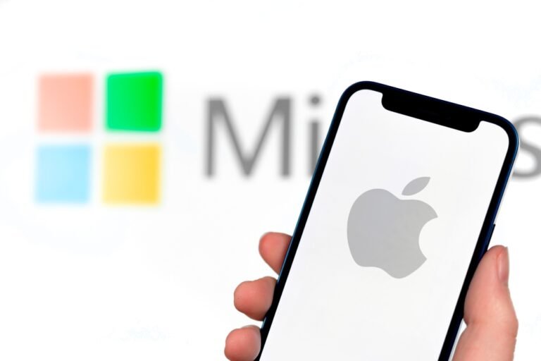 Apple telefon v ruce, v pozadí logo Microsoft. Apple vs. Microsoft